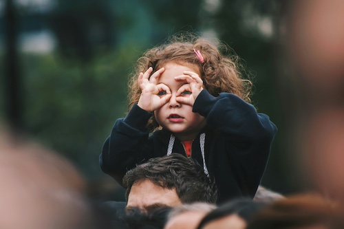 Ein Kind auf den Schultern eines Erwachsenen, beide Hände vor den Gesicht als Brille geformt.